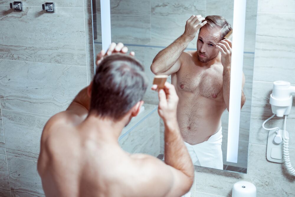 Athletic man. Athletic handsome bearded man combing his hair while standing in the bathroom