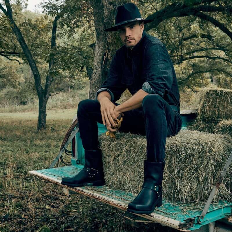 man wearing the frye Harness boots on top of bundled hay