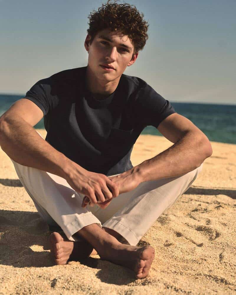 man sitting on the sand at a beach wearing black t-shirt and linen pants